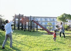 公園の滑り台の手前の芝生で親子が遊んでいる奥に3階建てのアパートが見える写真
