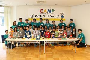 テーブルに並べられた創作品を前に記念撮影をしている緑色のTシャツを着たCAMPスタッフと大勢の子供たちの写真