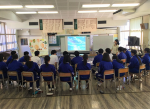 青いジャージを着た生徒たちが椅子に座り教室前方で流されている映像を見ている講義風景の写真