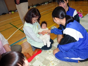 お母さんが座って抱っこしている赤ちゃんの手を握っている女子中学生の写真
