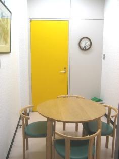 丸いテーブルを囲むように椅子が4脚あり、入口のドアが黄色の相談室の写真