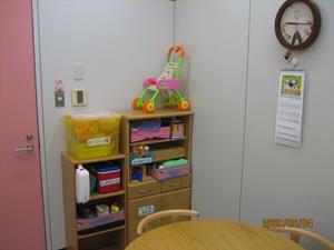 丸いテーブルに椅子が置いてあり、壁際には棚が置かれ、壁に時計とカレンダーが掛かり、入口のドアがピンク色の相談室の写真